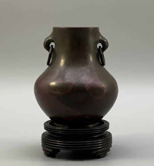 fig 6. A small pear-shaped dark bronze vase with loop handles and loose rings, featuring mottled red surface patches, resting on a carved dark wooden base.