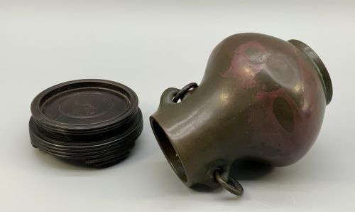 fig 7. A small pear-shaped dark bronze vase with loop handles and loose rings, featuring mottled red surface patches, resting on a carved dark wooden base.
