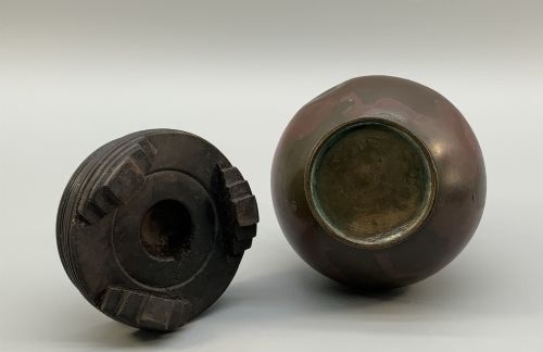 fig 8. A small pear-shaped dark bronze vase with loop handles and loose rings, featuring mottled red surface patches, resting on a carved dark wooden base.