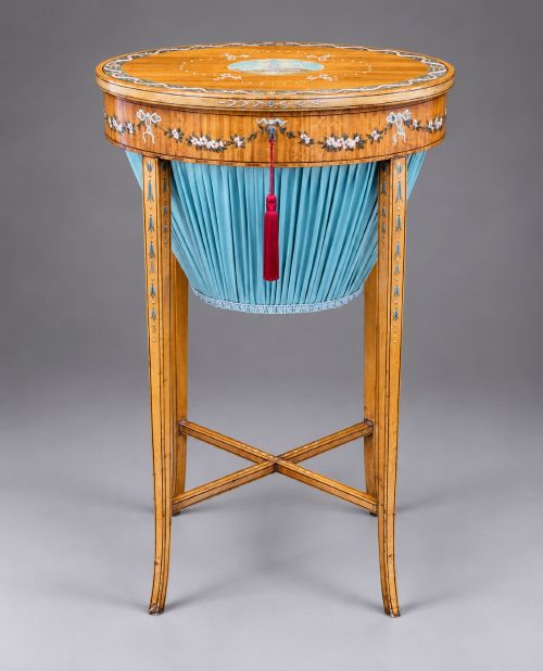fig 1. An oval satinwood sewing table featuring hand-painted floral swags, slender tapered legs, an X-form stretcher, and a pleated light blue fabric work basket suspended below.