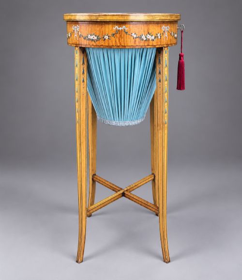 fig 2. An oval satinwood sewing table featuring hand-painted floral swags, slender tapered legs, an X-form stretcher, and a pleated light blue fabric work basket suspended below.