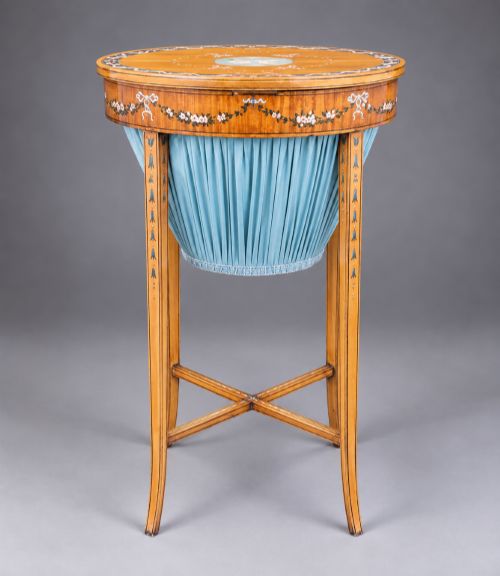 fig 3. An oval satinwood sewing table featuring hand-painted floral swags, slender tapered legs, an X-form stretcher, and a pleated light blue fabric work basket suspended below.