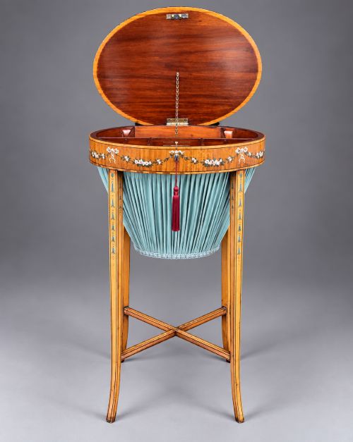 fig 4. An oval satinwood sewing table featuring hand-painted floral swags, slender tapered legs, an X-form stretcher, and a pleated light blue fabric work basket suspended below.