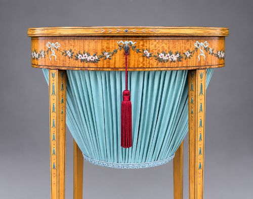 fig 5. An oval satinwood sewing table featuring hand-painted floral swags, slender tapered legs, an X-form stretcher, and a pleated light blue fabric work basket suspended below.