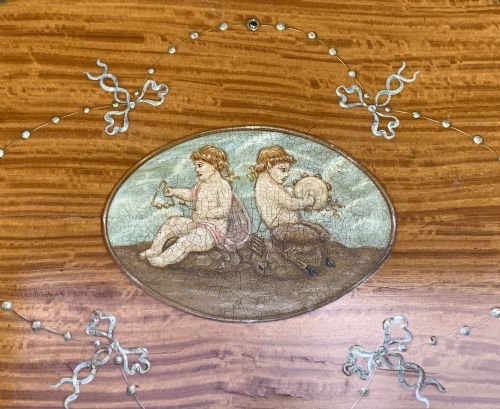 fig 8. An oval satinwood sewing table featuring hand-painted floral swags, slender tapered legs, an X-form stretcher, and a pleated light blue fabric work basket suspended below.