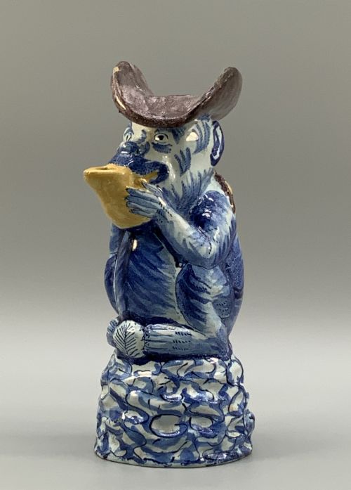fig 2. An 18th-century Dutch Delftware ceramic jug hand-painted in blue, white, brown, and yellow, designed in the form of a seated monkey in a tricorn hat holding a cup.