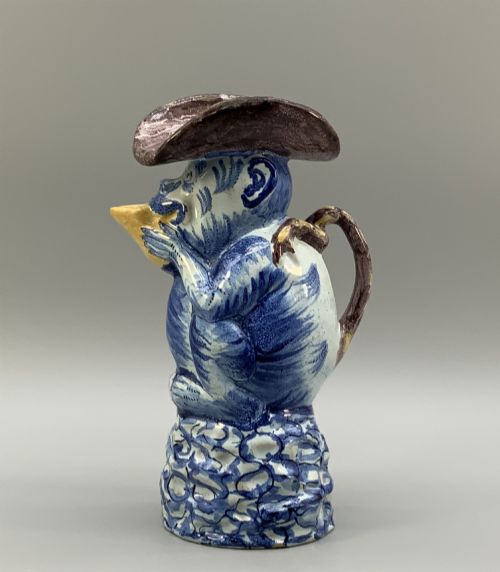 fig 3. An 18th-century Dutch Delftware ceramic jug hand-painted in blue, white, brown, and yellow, designed in the form of a seated monkey in a tricorn hat holding a cup.
