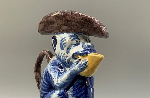 fig 5. An 18th-century Dutch Delftware ceramic jug hand-painted in blue, white, brown, and yellow, designed in the form of a seated monkey in a tricorn hat holding a cup.