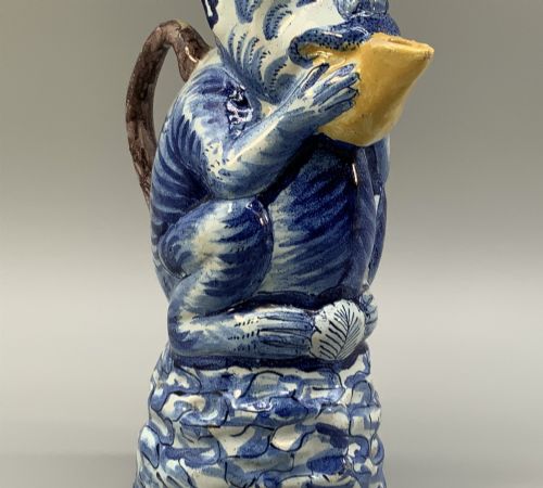 fig 6. An 18th-century Dutch Delftware ceramic jug hand-painted in blue, white, brown, and yellow, designed in the form of a seated monkey in a tricorn hat holding a cup.