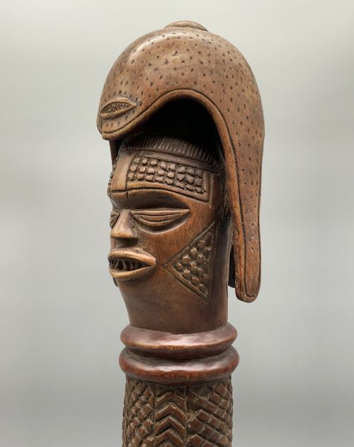 fig 7. A carved brown wooden African Mende Sowei staff finial depicting a stylised head with crest, mounted on a square black plinth.