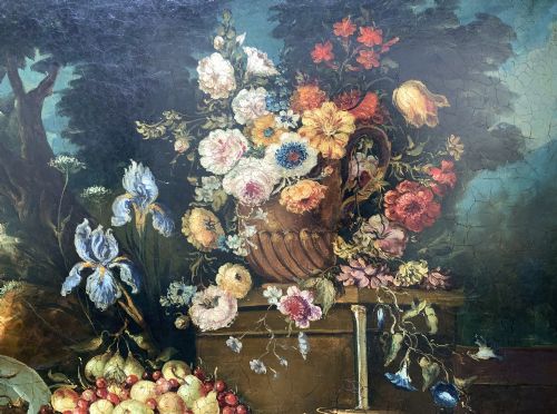 fig 5. A classical oil painting displaying a bowl of fruit, a urn with flowers, asparagus, and melons set against a dark landscape backdrop within a carved silver-grey frame.