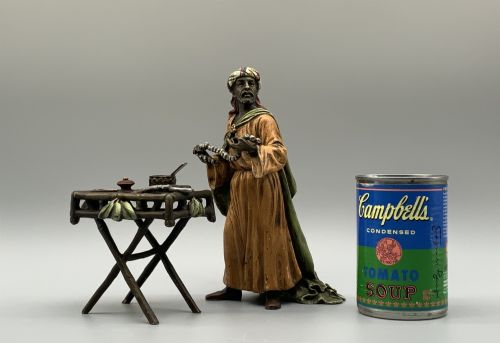 fig 1. A cold-painted bronze Orientalist figure wearing a turban and ochre robe, standing next to a small table displaying wares.