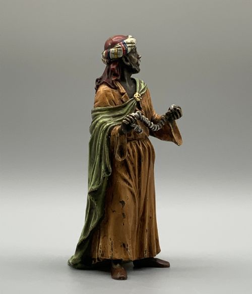 fig 6. A cold-painted bronze Orientalist figure wearing a turban and ochre robe, standing next to a small table displaying wares.