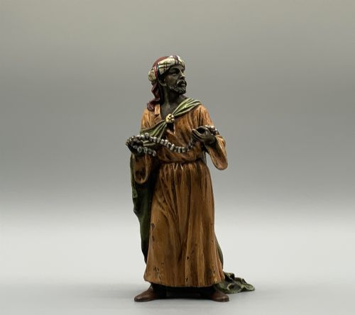 fig 7. A cold-painted bronze Orientalist figure wearing a turban and ochre robe, standing next to a small table displaying wares.