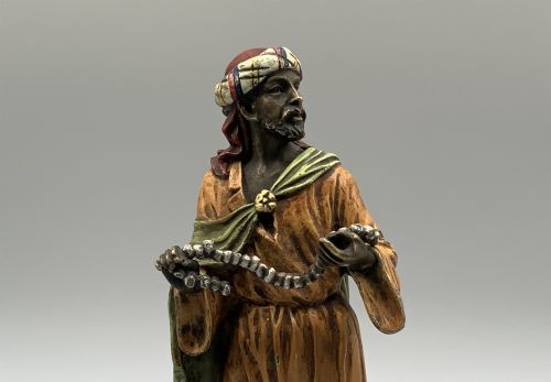 fig 8. A cold-painted bronze Orientalist figure wearing a turban and ochre robe, standing next to a small table displaying wares.