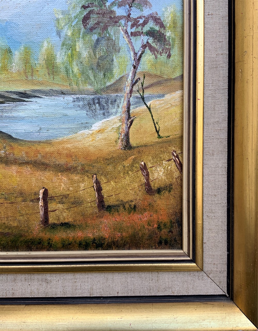 A mid-century oil landscape painting depicting tall gum trees beside a lake against blue mountains, framed in a gilt and fabric-lined wooden frame. View IX