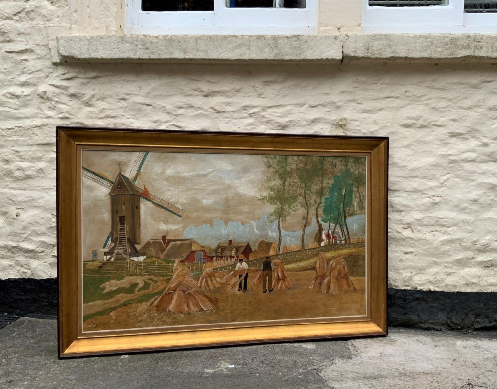 A mid-20th-century naive oil painting depicting agricultural workers gathering hay bales in a field beside a timber windmill, thatched cottages, and trees, presented in a gilt frame. View II