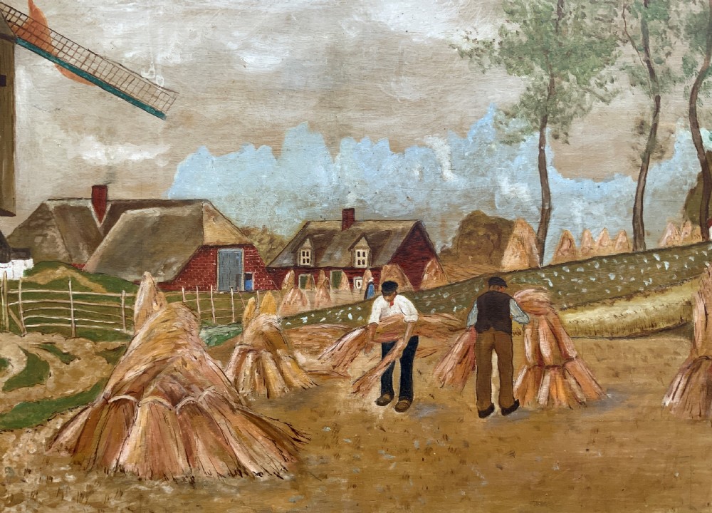 A mid-20th-century naive oil painting depicting agricultural workers gathering hay bales in a field beside a timber windmill, thatched cottages, and trees, presented in a gilt frame. View V