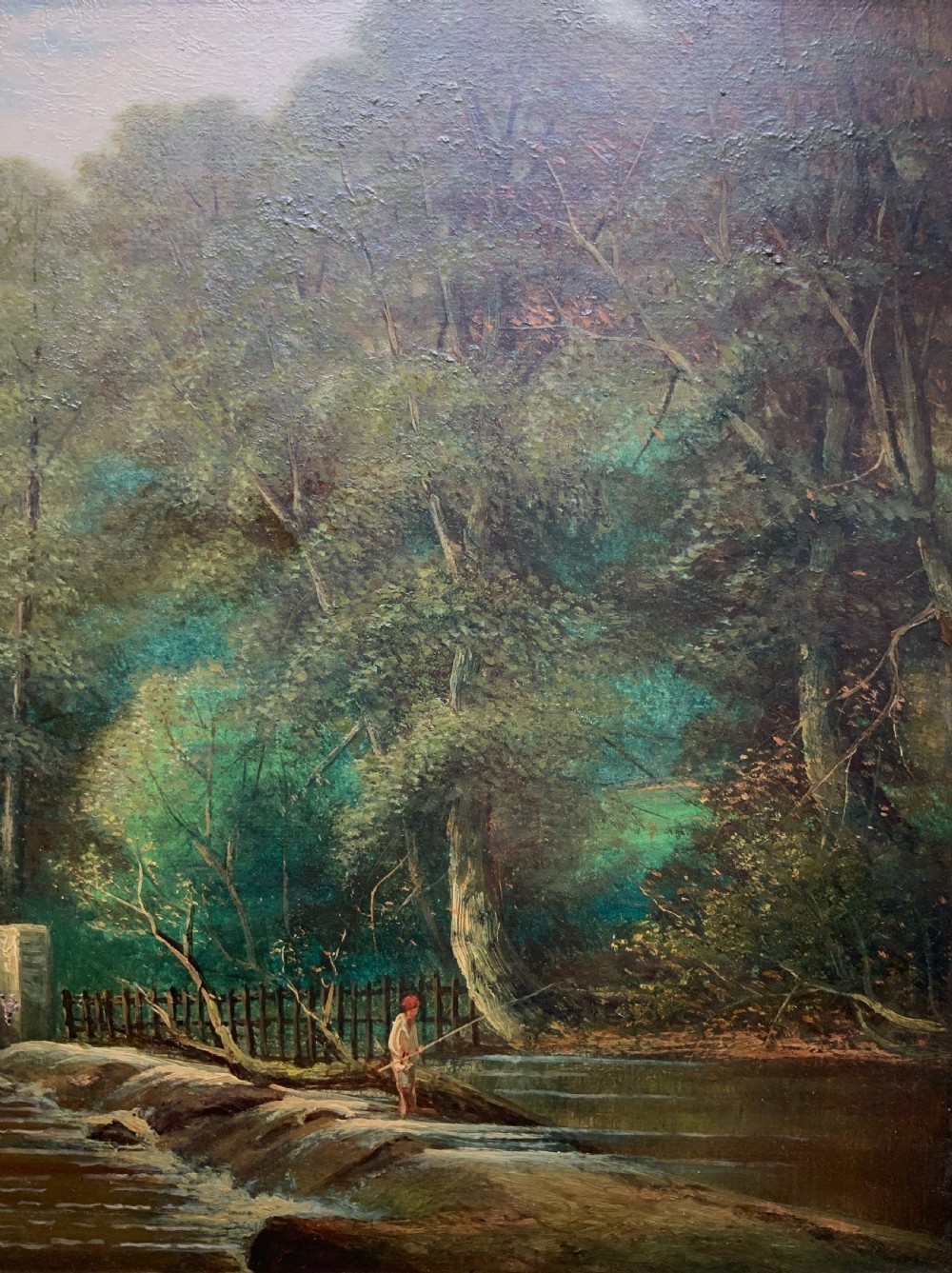 An early 20th-century landscape oil painting displaying a woodland river scene with a figure fishing near a weir, encased in a deep gold frame. View III