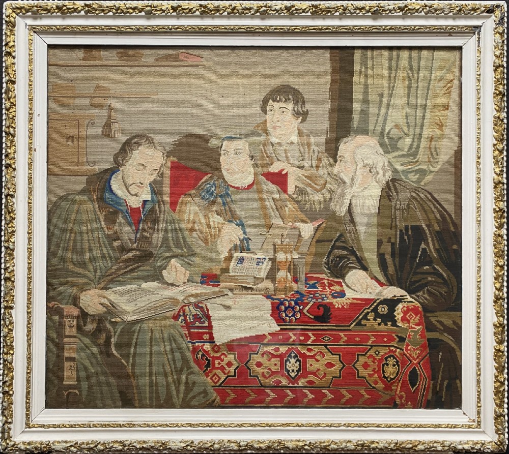 A framed mid-nineteenth-century Berlin woolwork tapestry depicting four historical figures seated around a carpet-covered table, housed within a gilt decorative frame.
