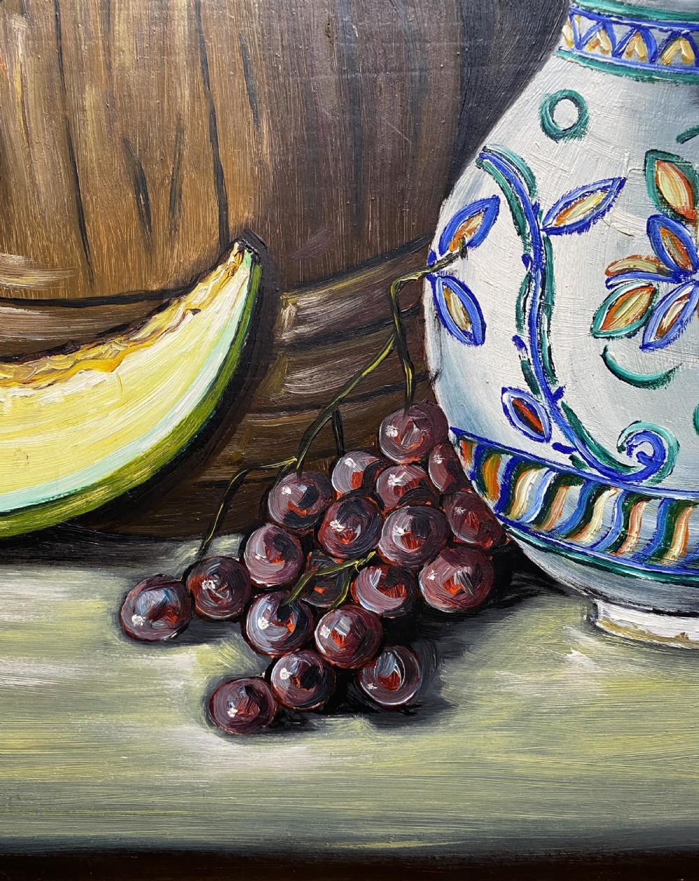 A framed mid-century oil painting depicting a still life composition with a blue patterned ceramic jug, a wooden barrel, a melon slice, and purple grapes. View IV