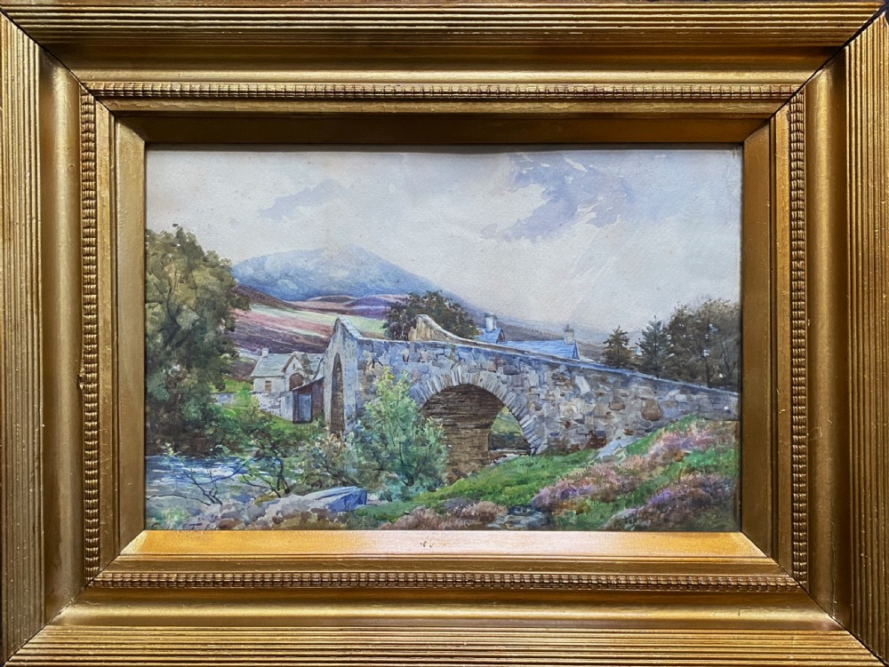 An early 20th-century watercolour painting depicting a stone arch bridge over a river surrounded by hills, set within a wide gold frame.