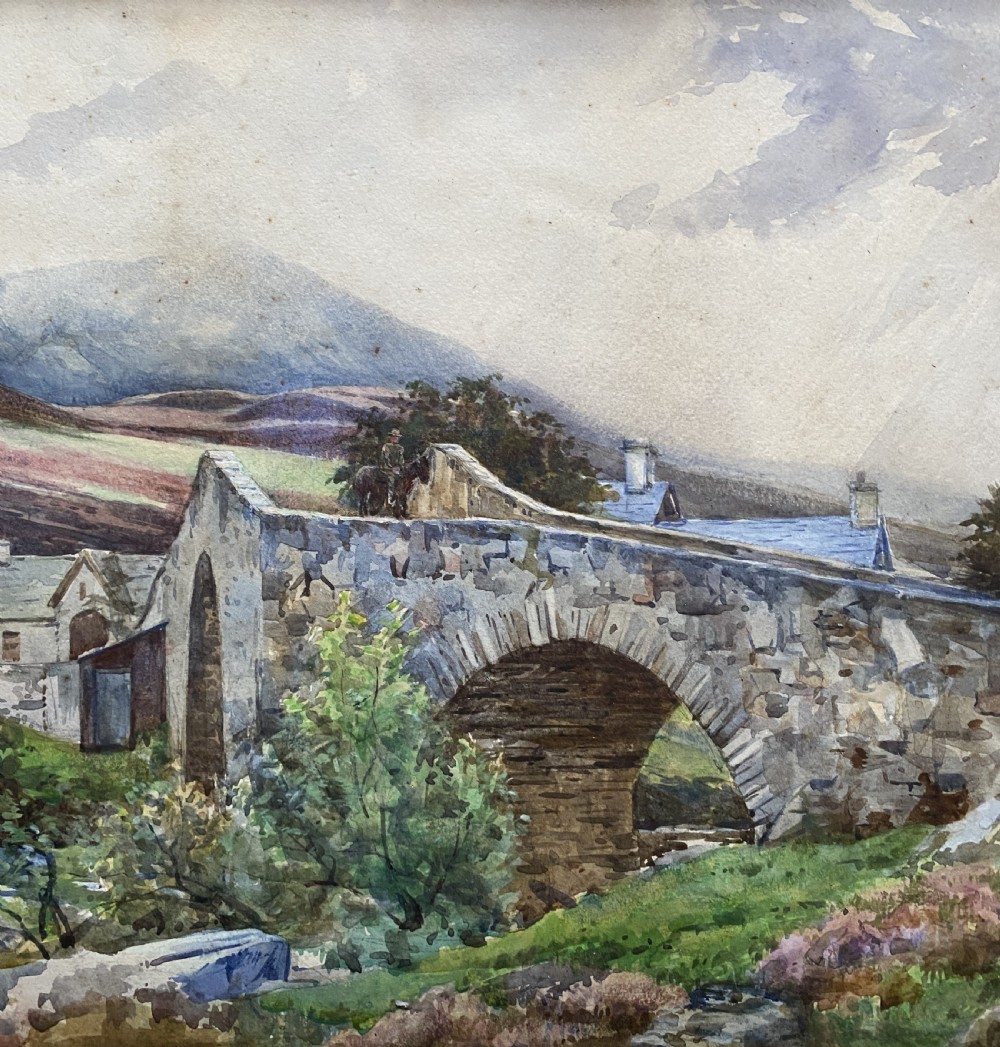 An early 20th-century watercolour painting depicting a stone arch bridge over a river surrounded by hills, set within a wide gold frame. View IV