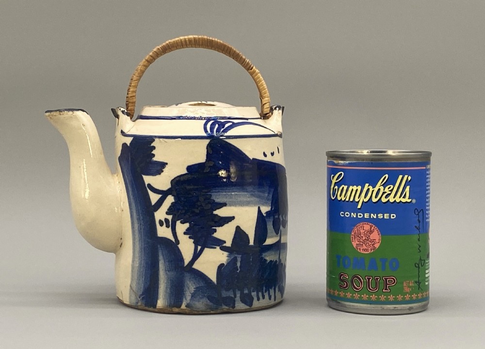 A cylindrical cream-coloured ceramic teapot decorated with hand-painted blue cobalt landscapes and featuring an overhead woven cane handle. View II