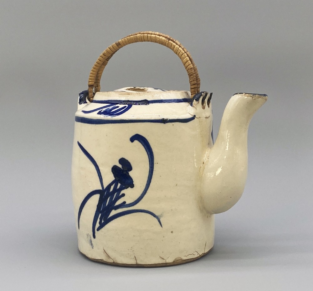 A cylindrical cream-coloured ceramic teapot decorated with hand-painted blue cobalt landscapes and featuring an overhead woven cane handle. View III