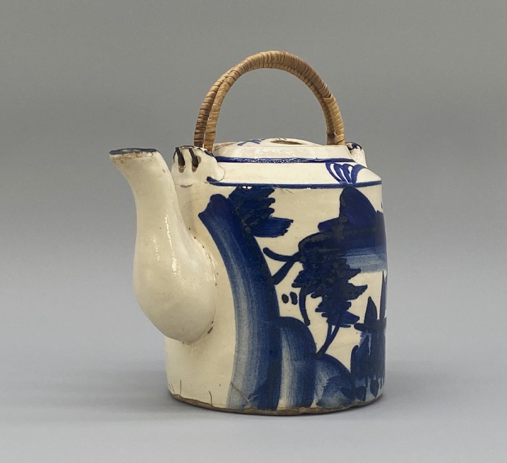 A cylindrical cream-coloured ceramic teapot decorated with hand-painted blue cobalt landscapes and featuring an overhead woven cane handle. View V