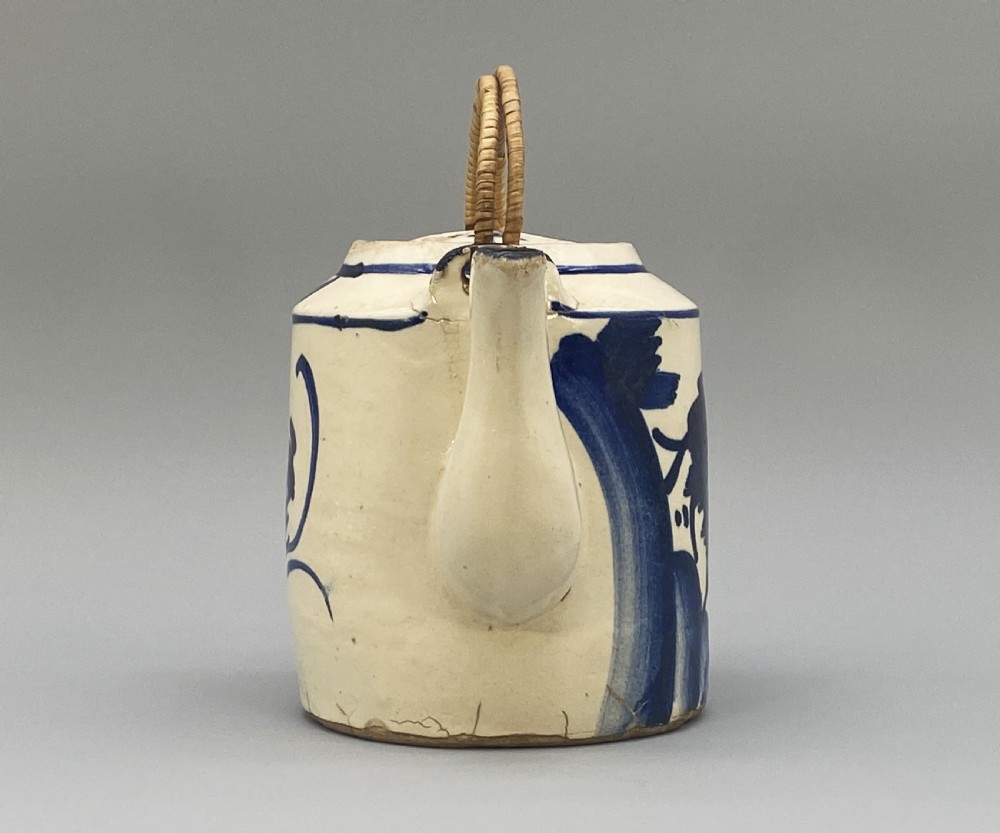 A cylindrical cream-coloured ceramic teapot decorated with hand-painted blue cobalt landscapes and featuring an overhead woven cane handle. View VI