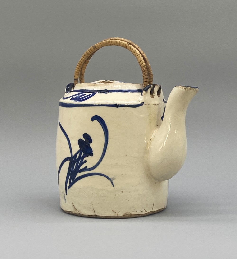 A cylindrical cream-coloured ceramic teapot decorated with hand-painted blue cobalt landscapes and featuring an overhead woven cane handle. View VII