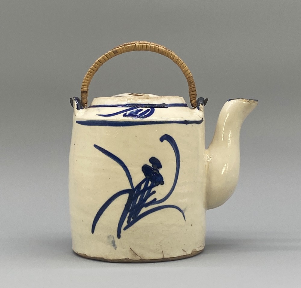 A cylindrical cream-coloured ceramic teapot decorated with hand-painted blue cobalt landscapes and featuring an overhead woven cane handle. View VIII