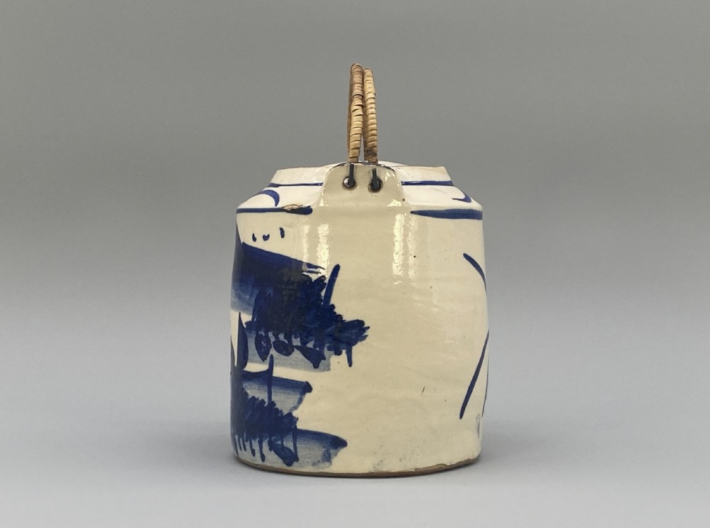 A cylindrical cream-coloured ceramic teapot decorated with hand-painted blue cobalt landscapes and featuring an overhead woven cane handle. View IX