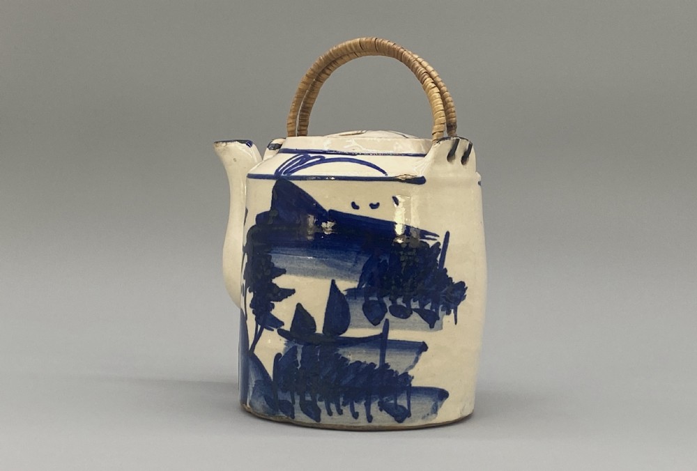 A cylindrical cream-coloured ceramic teapot decorated with hand-painted blue cobalt landscapes and featuring an overhead woven cane handle. View X