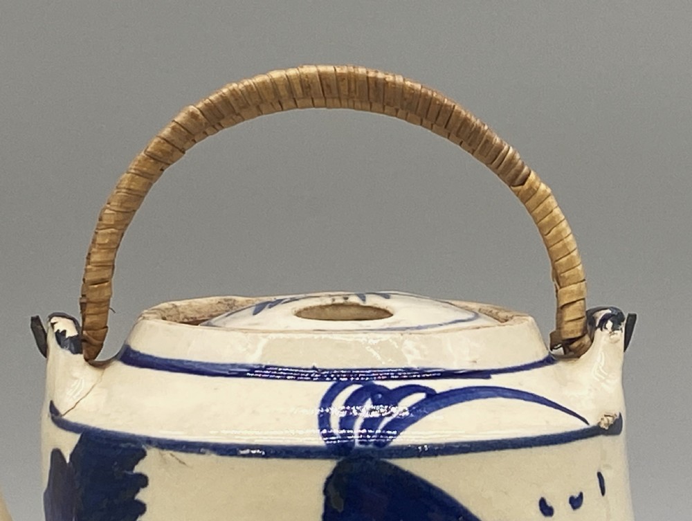 A cylindrical cream-coloured ceramic teapot decorated with hand-painted blue cobalt landscapes and featuring an overhead woven cane handle. View XI