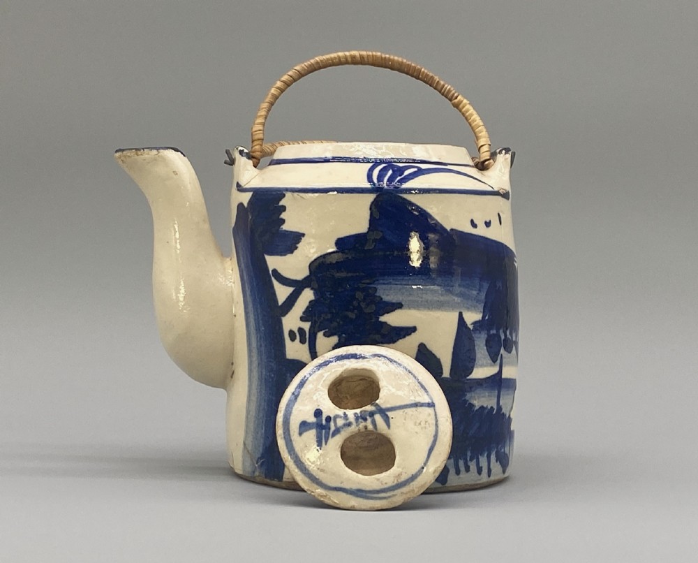 A cylindrical cream-coloured ceramic teapot decorated with hand-painted blue cobalt landscapes and featuring an overhead woven cane handle. View XII
