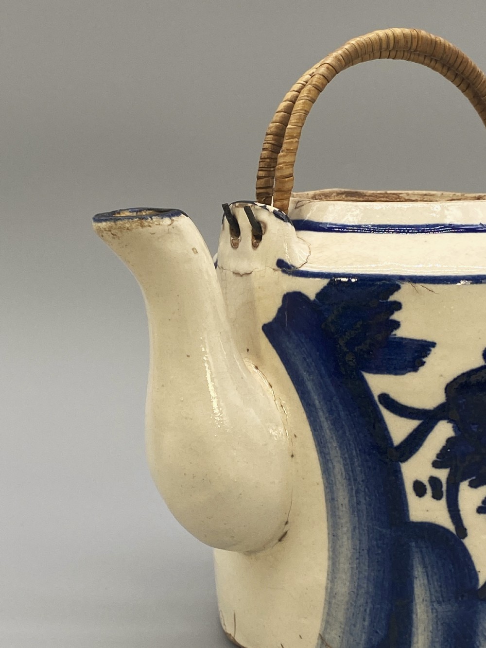 A cylindrical cream-coloured ceramic teapot decorated with hand-painted blue cobalt landscapes and featuring an overhead woven cane handle. View XV