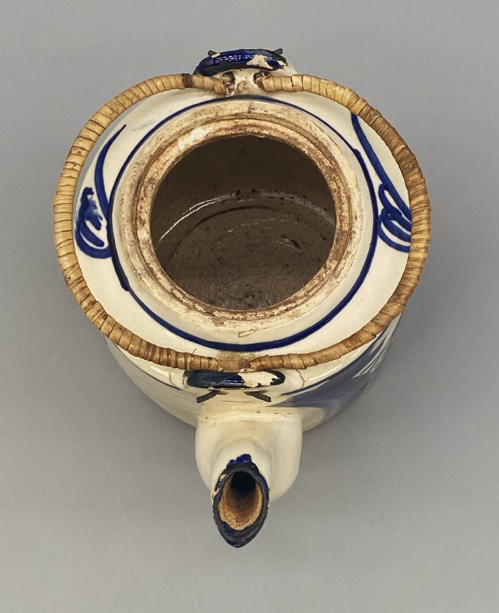 A cylindrical cream-coloured ceramic teapot decorated with hand-painted blue cobalt landscapes and featuring an overhead woven cane handle. View XVI