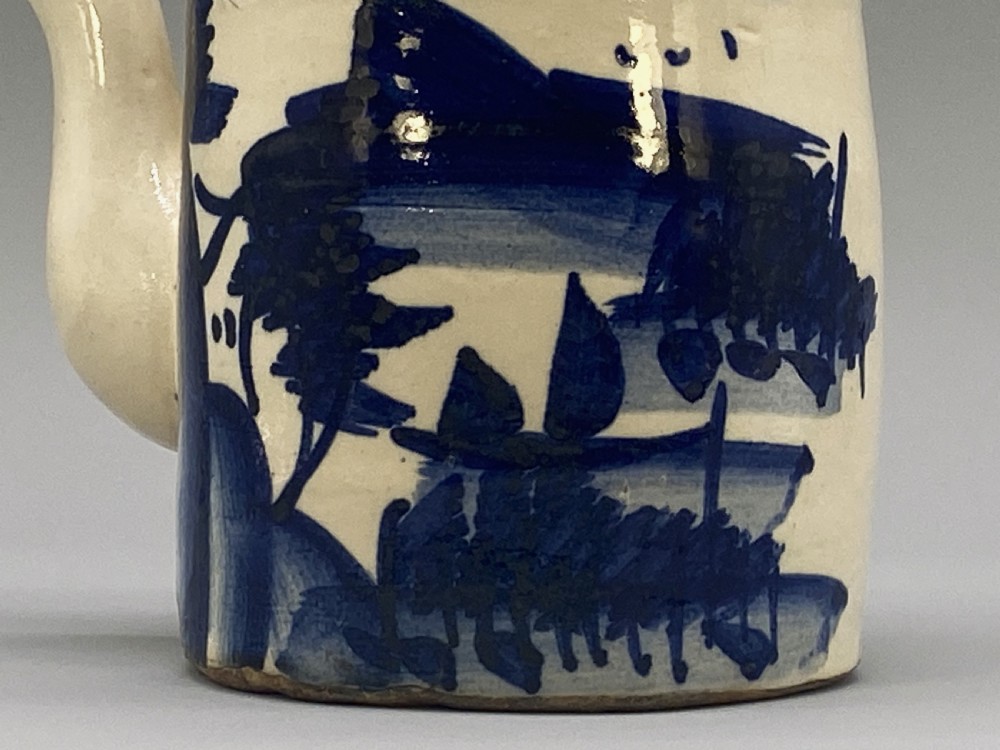 A cylindrical cream-coloured ceramic teapot decorated with hand-painted blue cobalt landscapes and featuring an overhead woven cane handle. View XVIII
