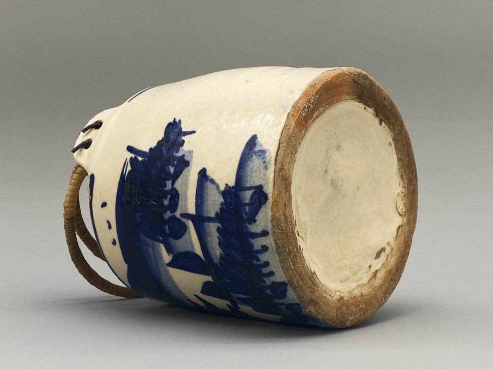 A cylindrical cream-coloured ceramic teapot decorated with hand-painted blue cobalt landscapes and featuring an overhead woven cane handle. View XIX