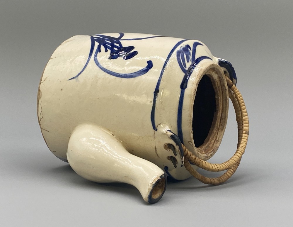 A cylindrical cream-coloured ceramic teapot decorated with hand-painted blue cobalt landscapes and featuring an overhead woven cane handle. View XX