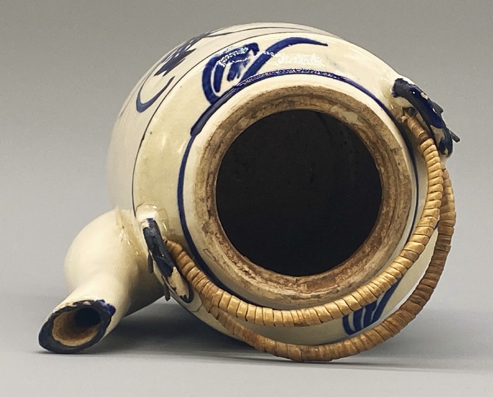 A cylindrical cream-coloured ceramic teapot decorated with hand-painted blue cobalt landscapes and featuring an overhead woven cane handle. View XXI