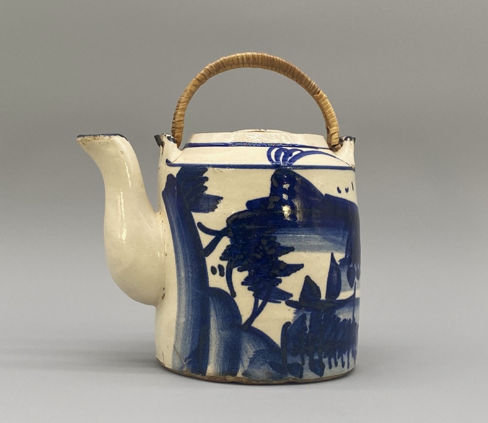 A cylindrical cream-coloured ceramic teapot decorated with hand-painted blue cobalt landscapes and featuring an overhead woven cane handle. View XXII