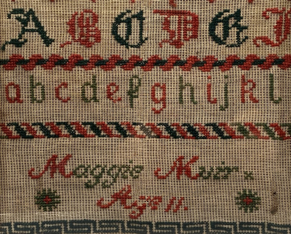 A vertical Victorian framed sampler featuring rows of embroidered alphabets in red and green wool thread on linen fabric, housed within a gilded wooden frame. View IV