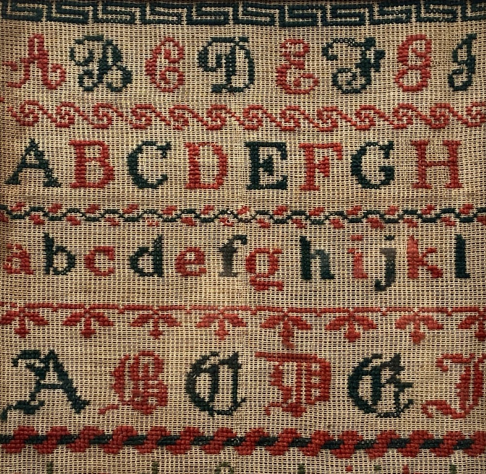 A vertical Victorian framed sampler featuring rows of embroidered alphabets in red and green wool thread on linen fabric, housed within a gilded wooden frame. View V