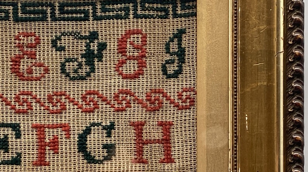 A vertical Victorian framed sampler featuring rows of embroidered alphabets in red and green wool thread on linen fabric, housed within a gilded wooden frame. View VI