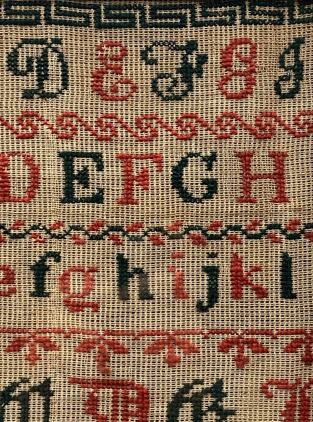 A vertical Victorian framed sampler featuring rows of embroidered alphabets in red and green wool thread on linen fabric, housed within a gilded wooden frame. View VII
