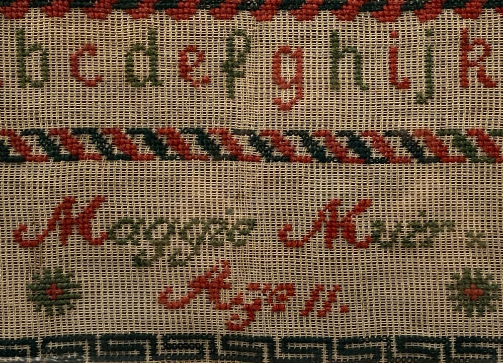 A vertical Victorian framed sampler featuring rows of embroidered alphabets in red and green wool thread on linen fabric, housed within a gilded wooden frame. View VIII