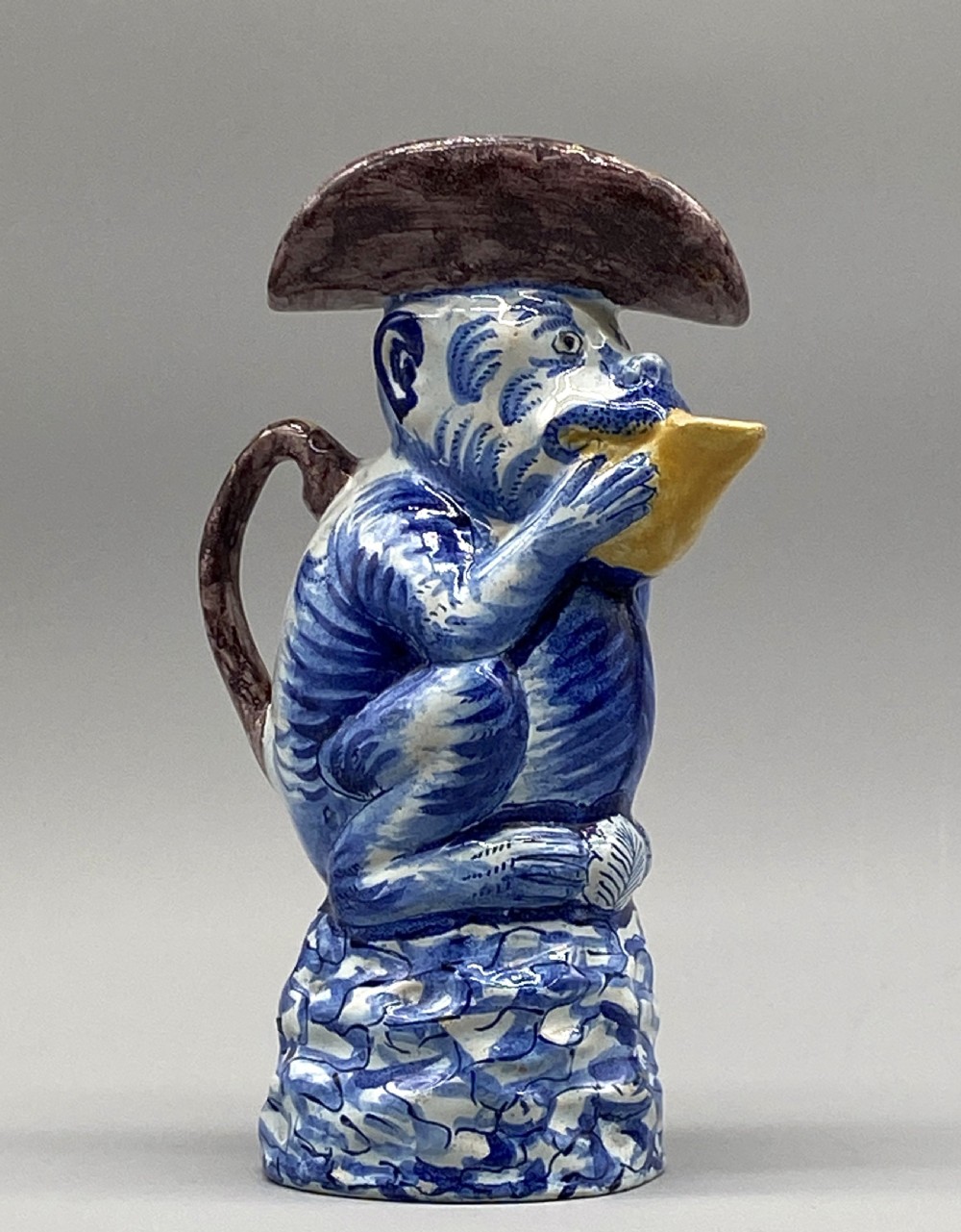 An eighteenth-century Dutch Delft tin-glazed earthenware jug shaped as a seated monkey wearing a hat and holding a vessel, decorated in blue, white, brown, and yellow glazes.